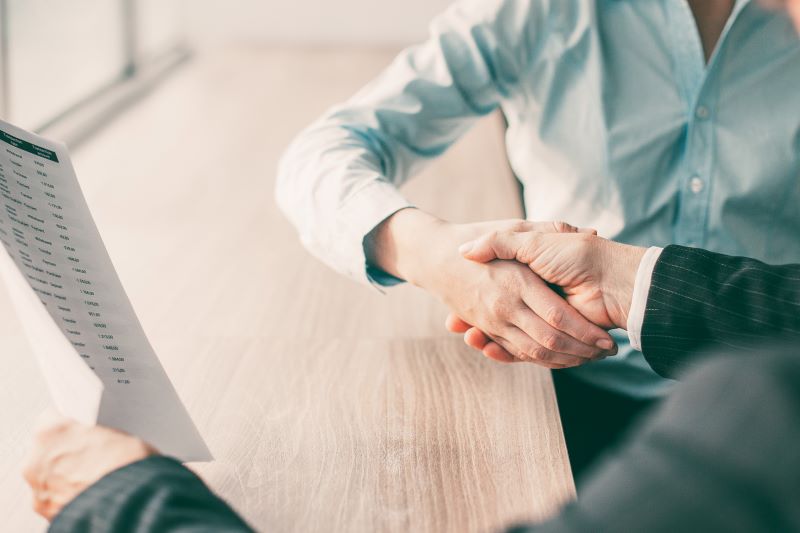 Stretta di mano tra due persone in un concessionario con un'auto bianca sullo sfondo, simbolo di un accordo per l'uso promiscuo dell'auto.